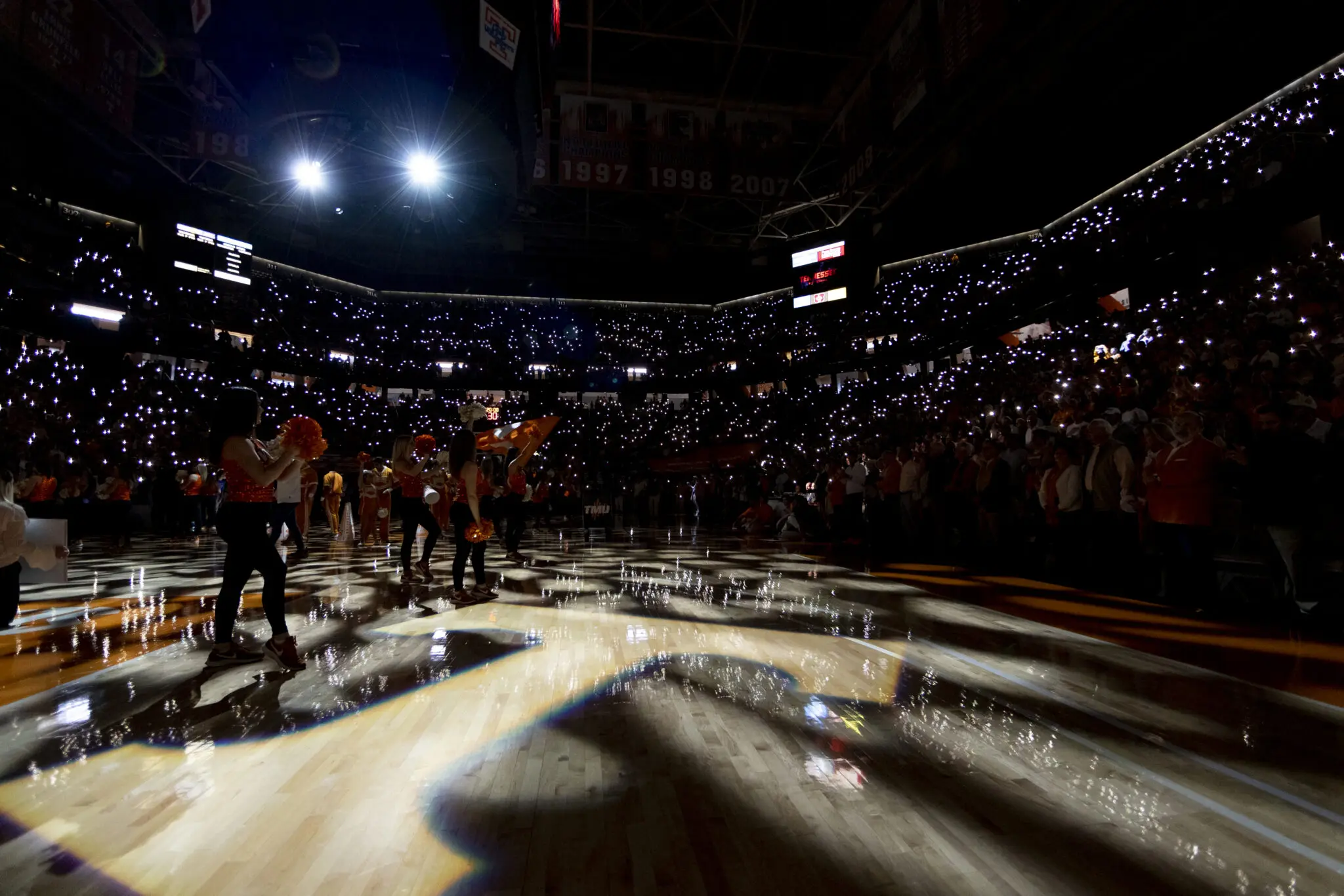 Basketball players stand on a dimly lit court with bright arena lights above.
