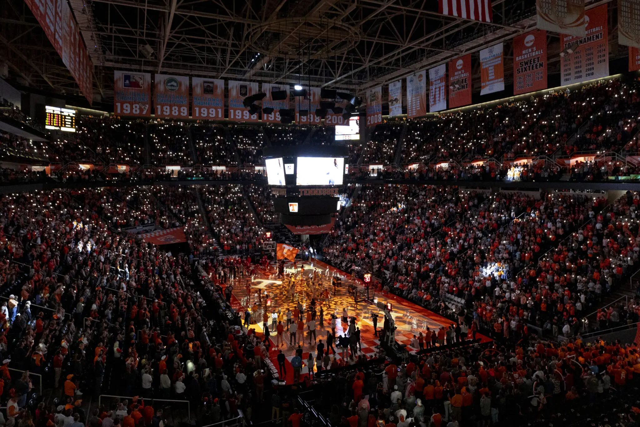 A packed indoor basketball arena during a game with bright court lights.
