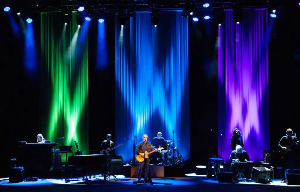 A band performs on stage with colorful blue, green, and purple lighting.