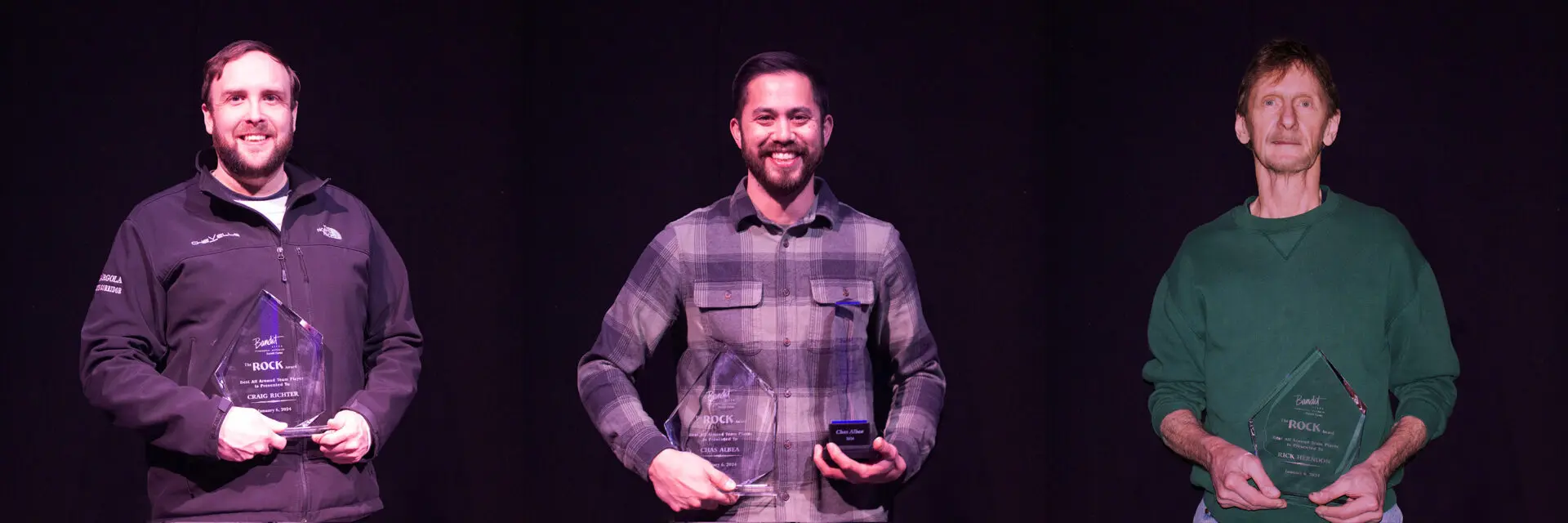 Smiling man in a plaid shirt holding a microphone on stage.