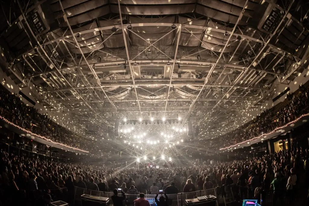 A packed arena shines under bright stage lights during a live concert.