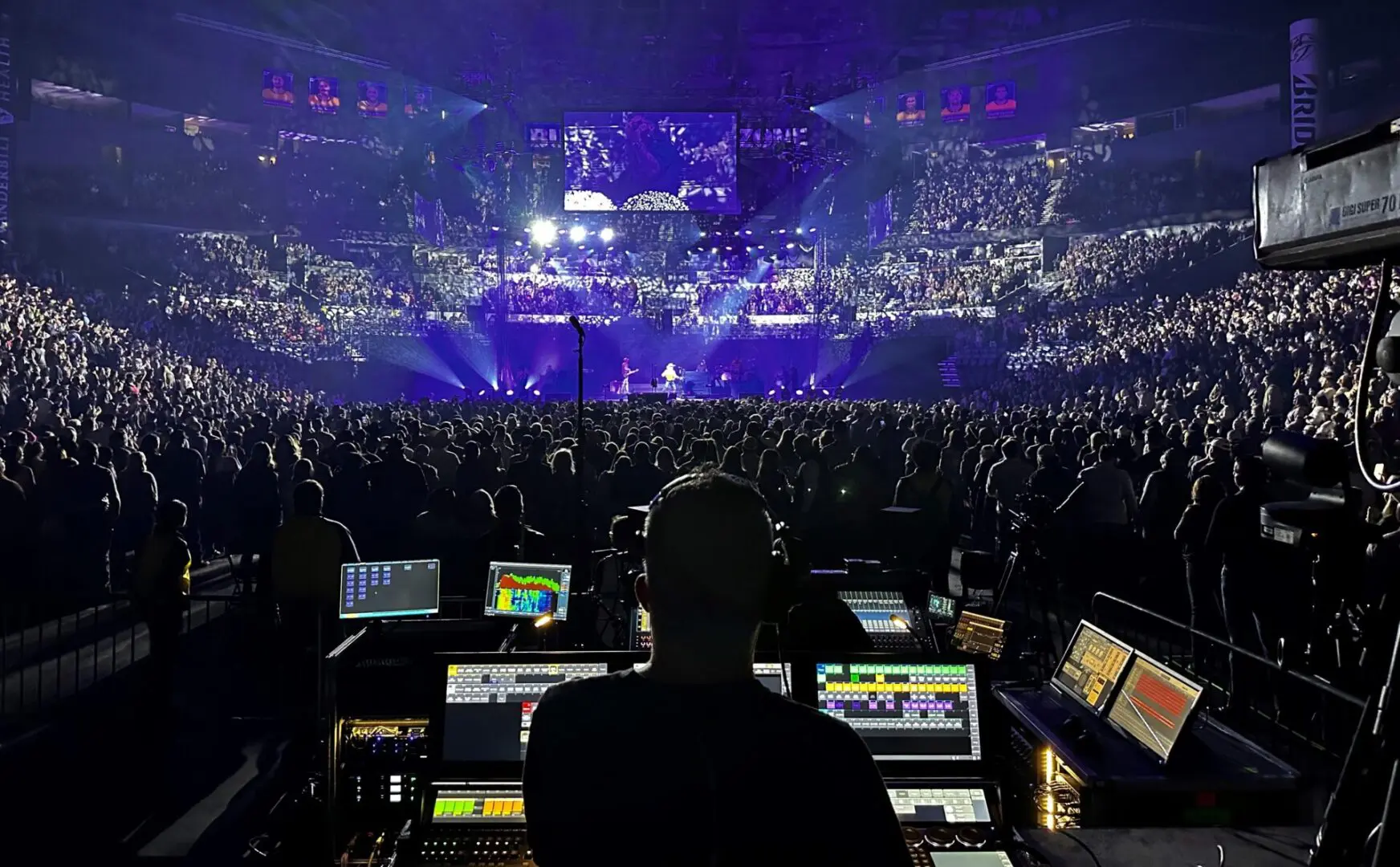 A sound engineer manages audio at a packed indoor concert with vibrant purple lighting.