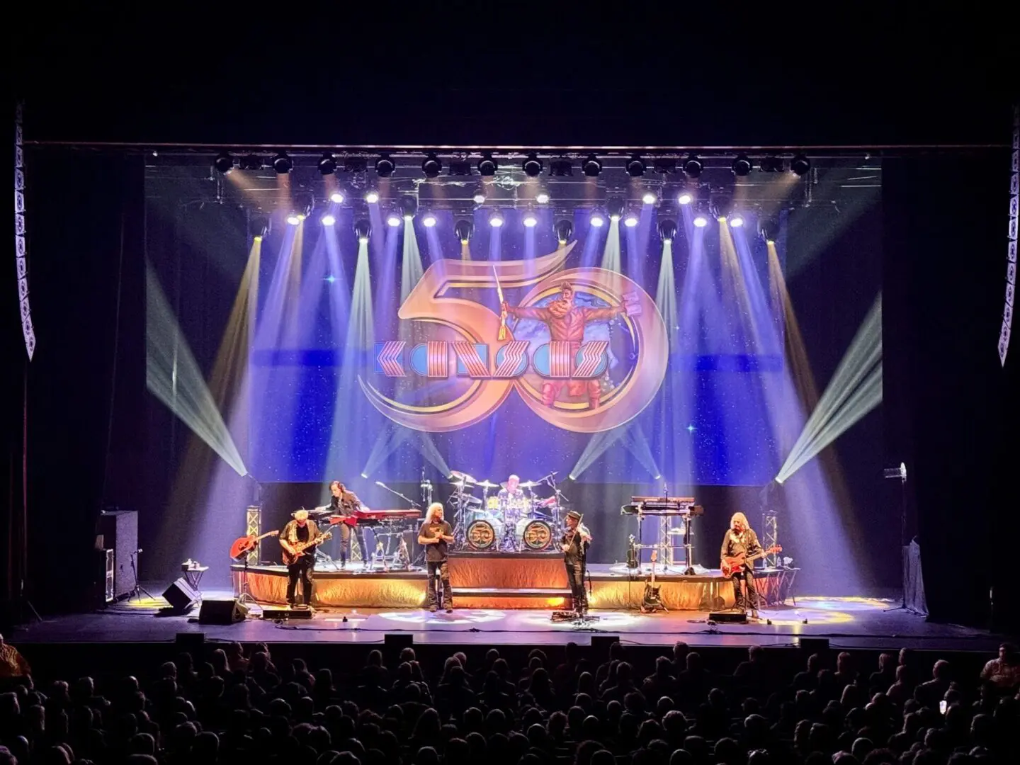 A live band performs on stage with vibrant lighting and a large logo backdrop.