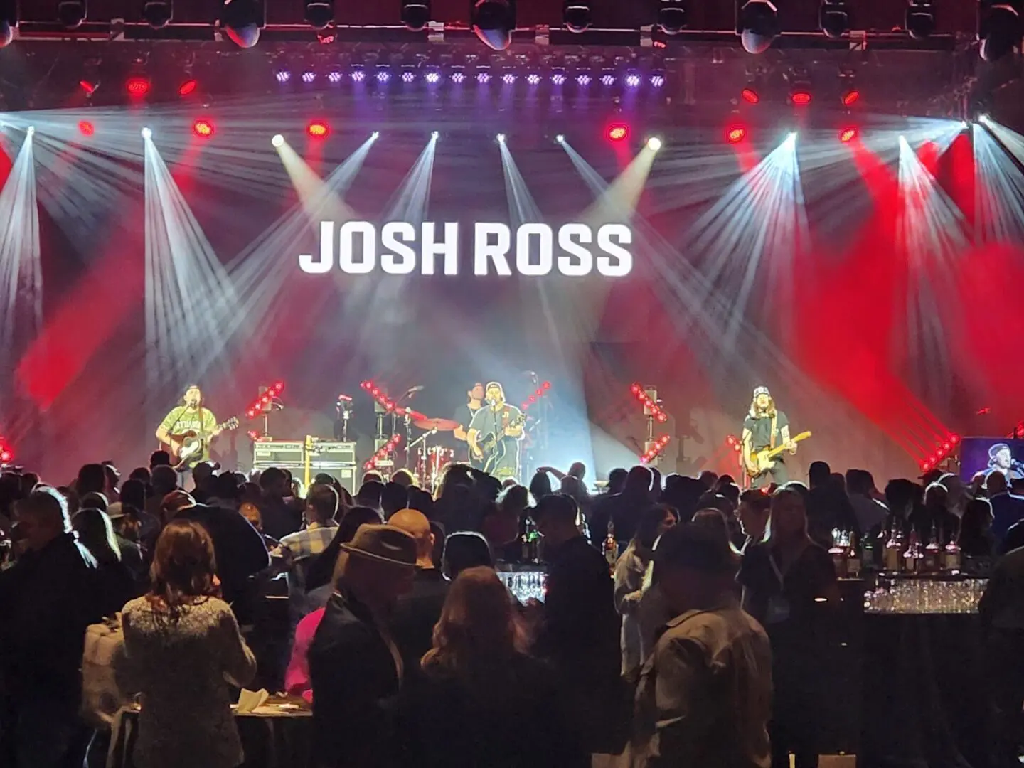 A live band performs on stage with bright spotlights and a crowd watching.