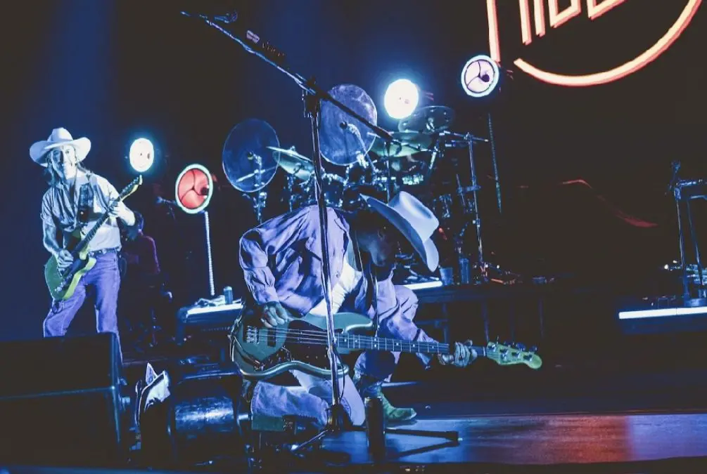 Musician playing guitar and singing on stage with drum set behind.