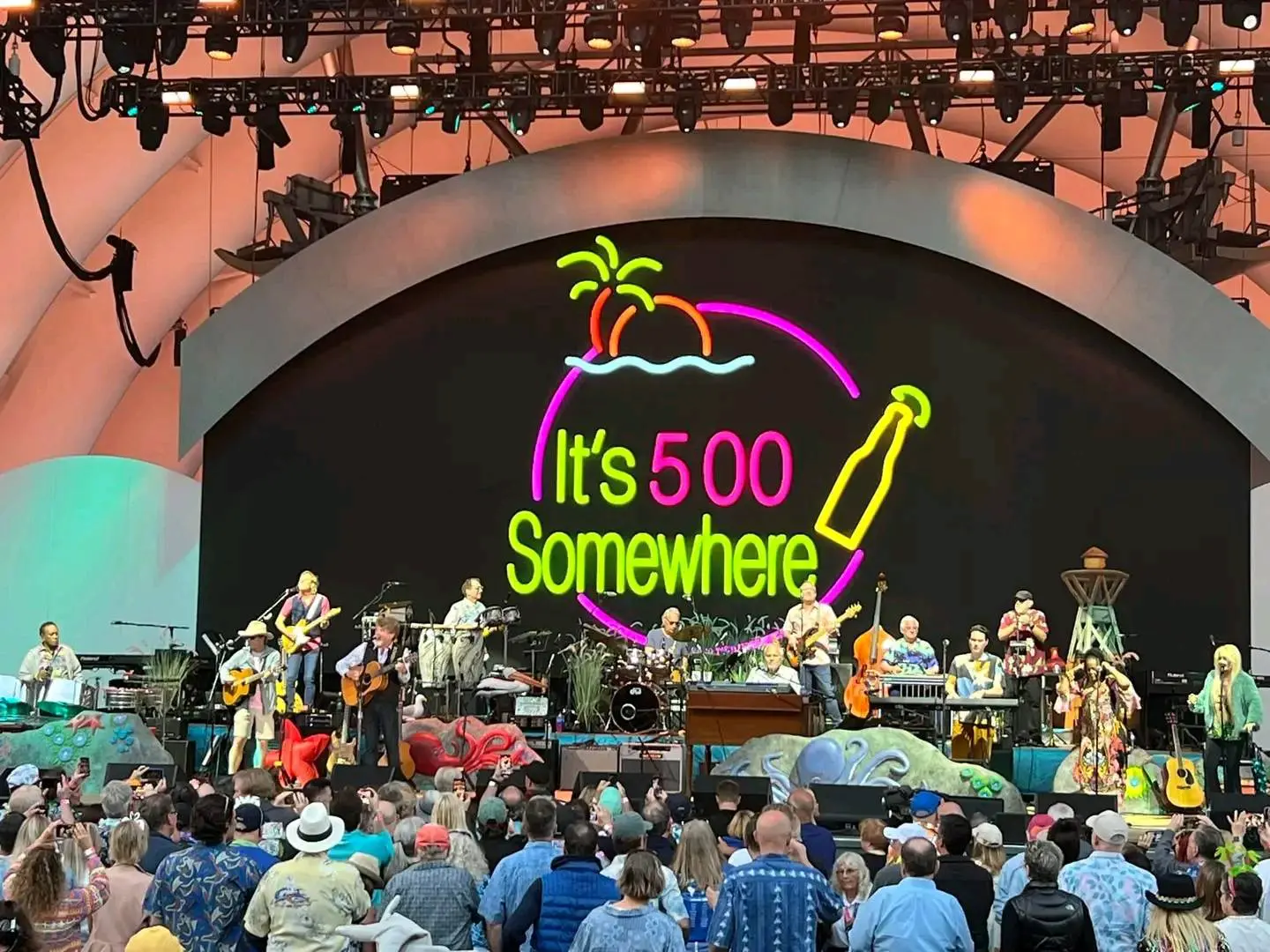 Outdoor concert with a large crowd at a purple-lit band shell.