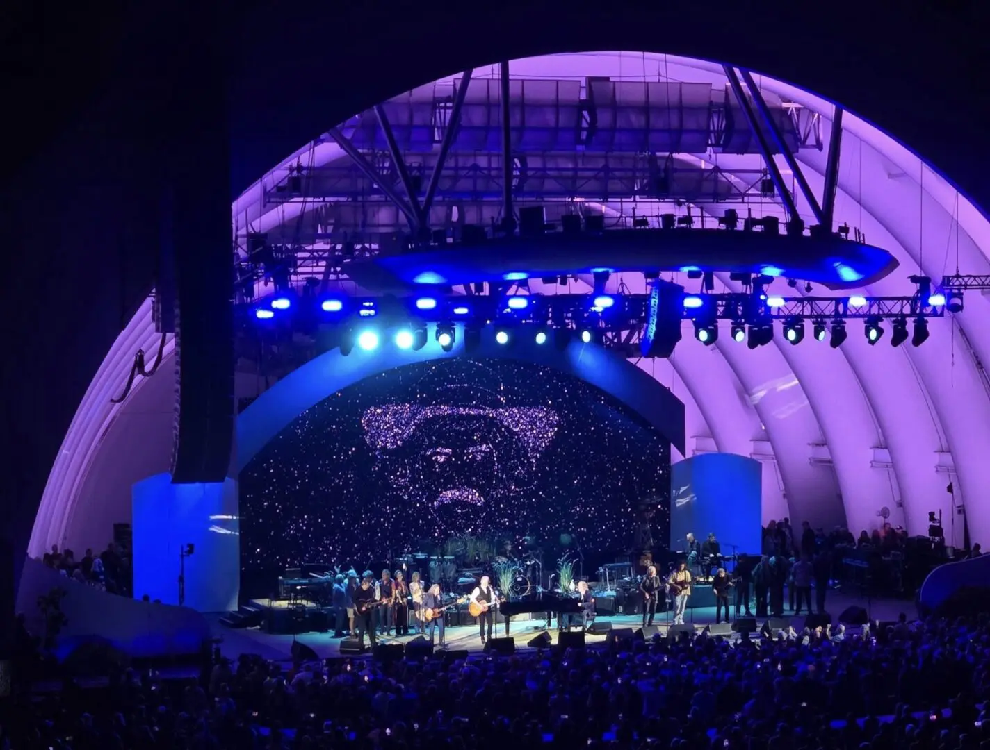 Outdoor concert with a large crowd at a purple-lit band shell.