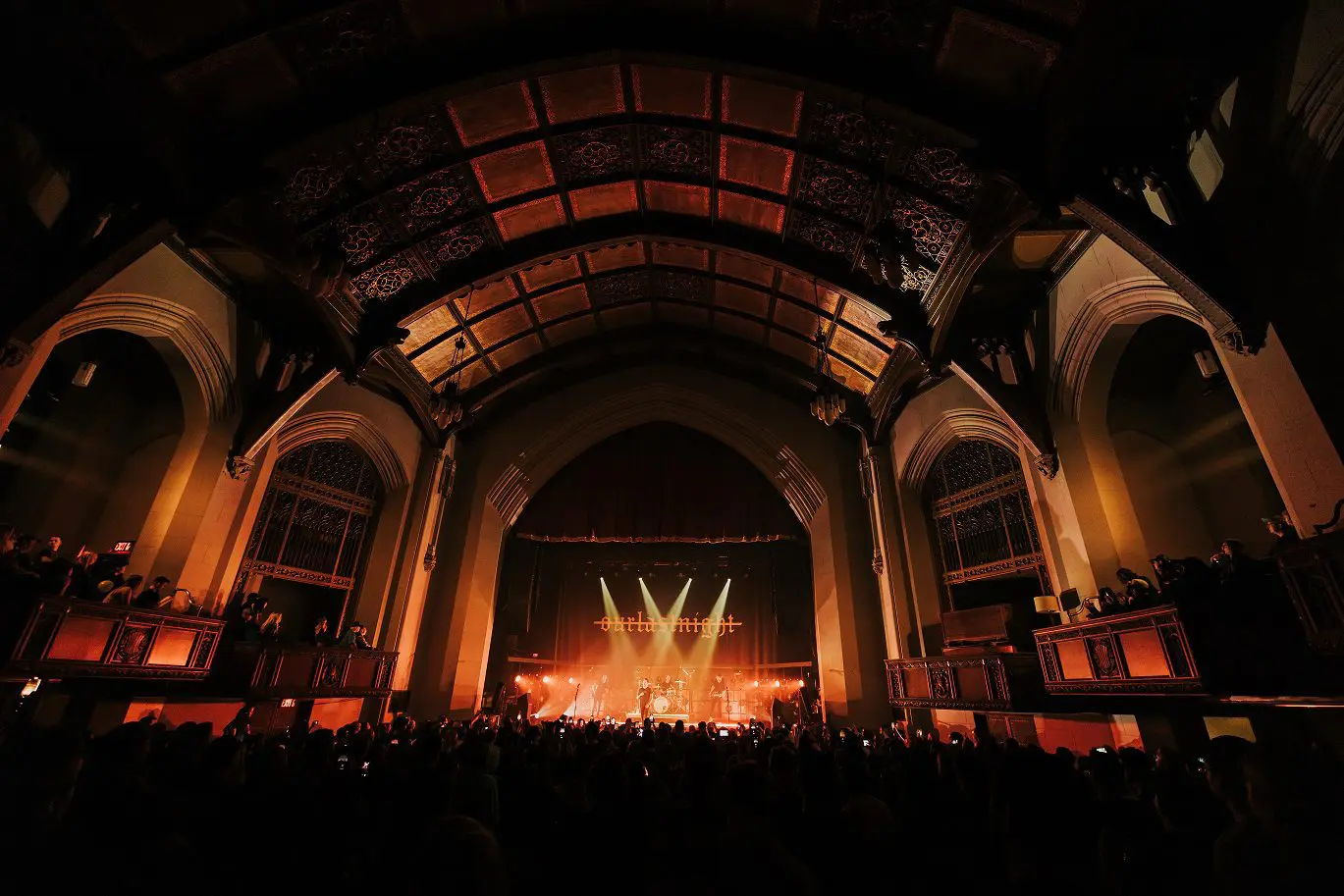 A grand concert with bright stage lights in a large, arched venue filled with an audience.