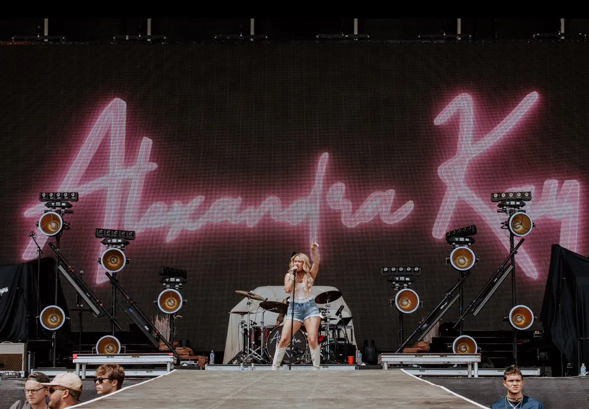 DJ Alexander R performing at an outdoor event with a neon sign.
