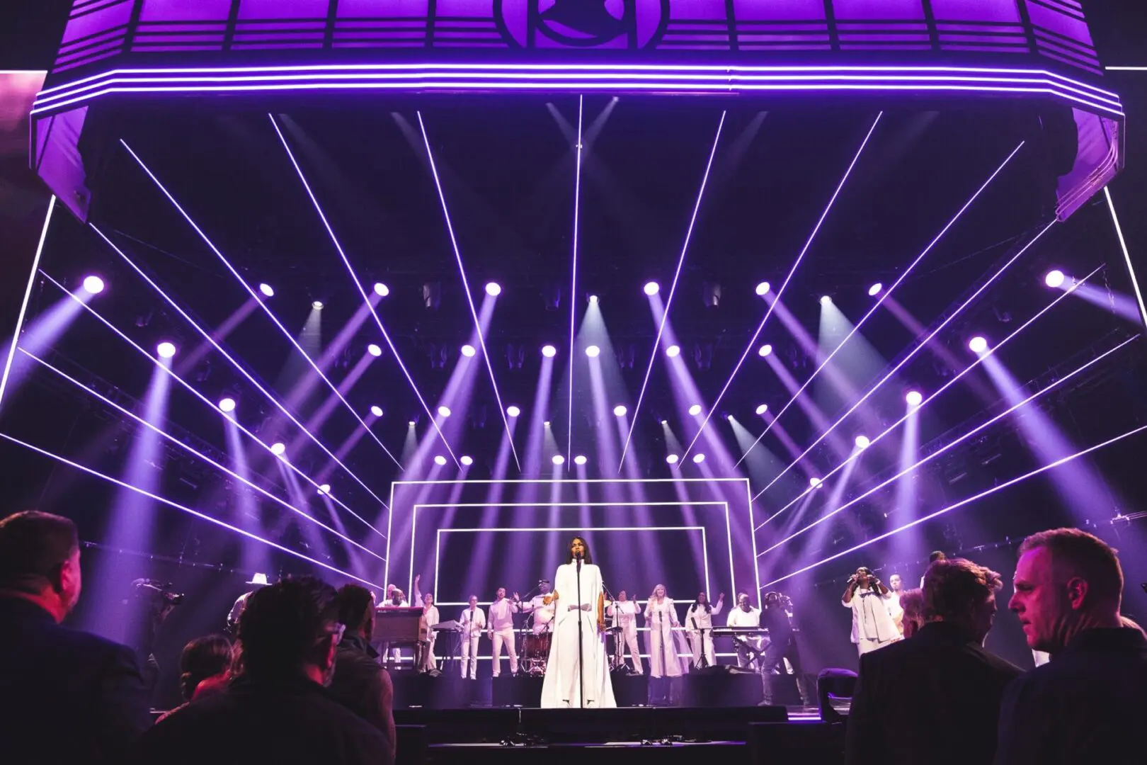 A singer performs on stage with a choir and purple lights.