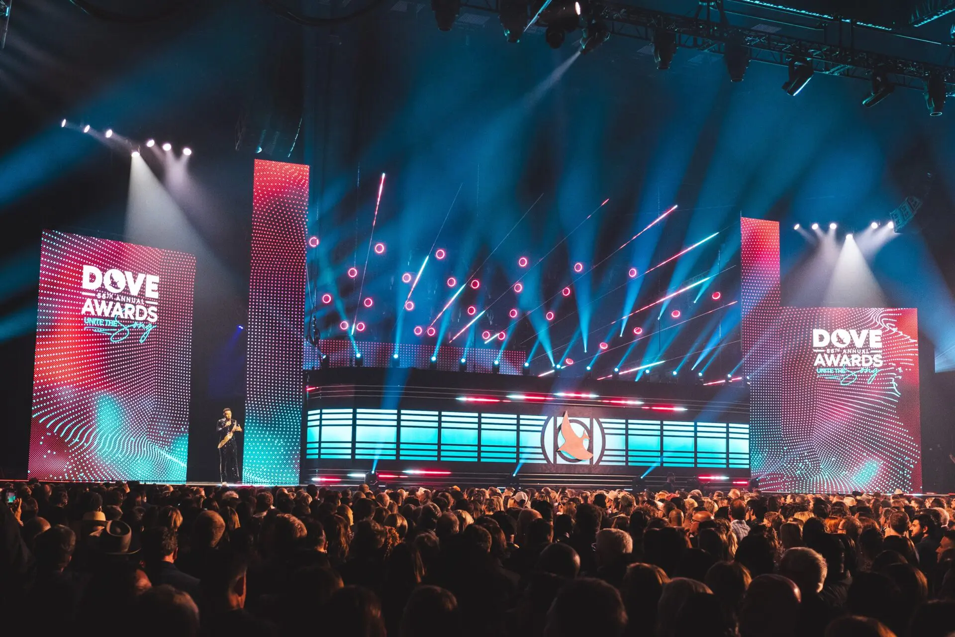 A large crowd watches a vibrant light show on a stage with illuminated screens.