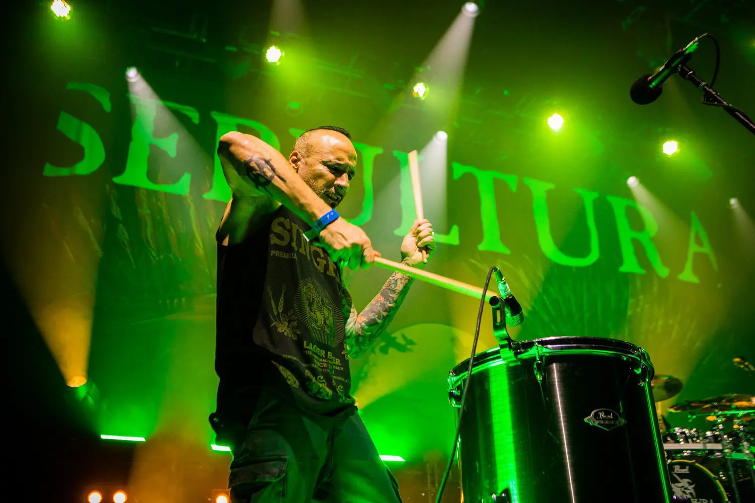 A drummer passionately playing drums on stage with green lighting.