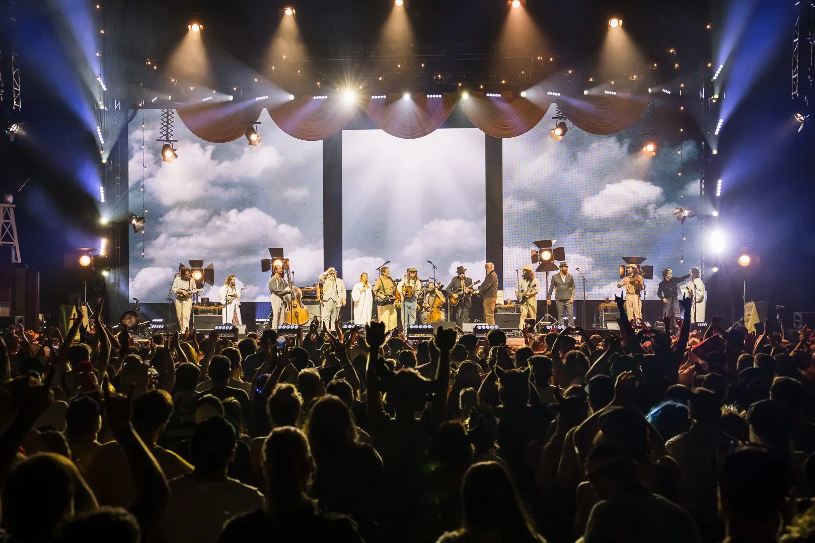 A large band performs on stage with dramatic sky visuals behind them.