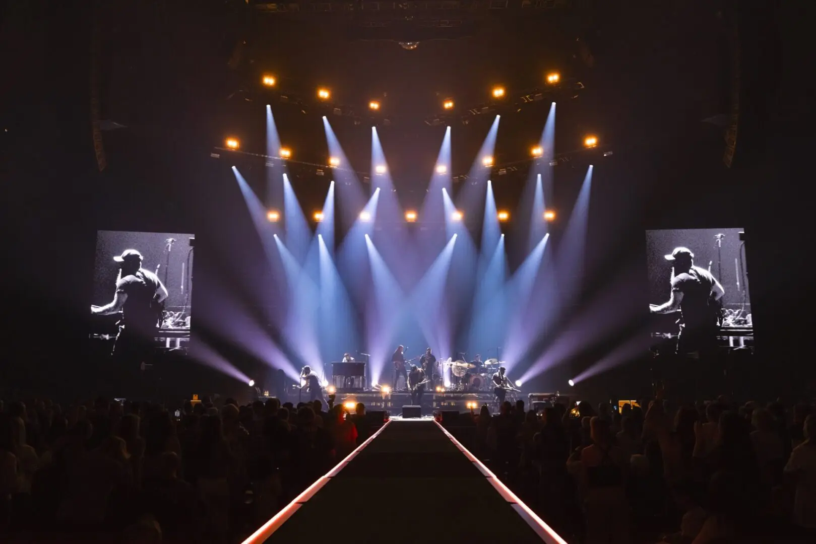 A band performs on a brightly lit stage with large screens and an enthusiastic audience.