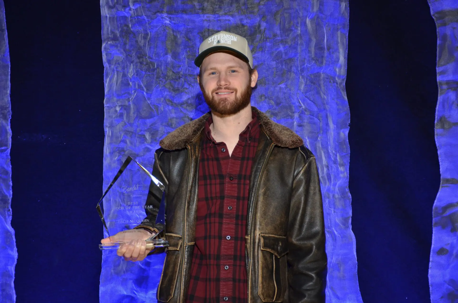 Man in a leather jacket and cap holding an award on stage.