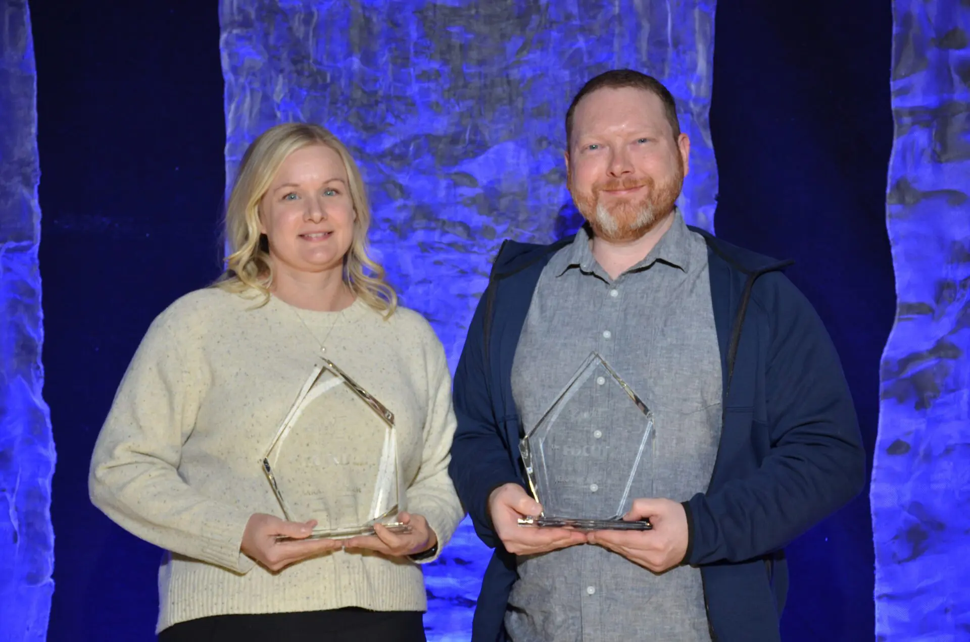 Two people holding awards, smiling at a formal event with blue lighting.