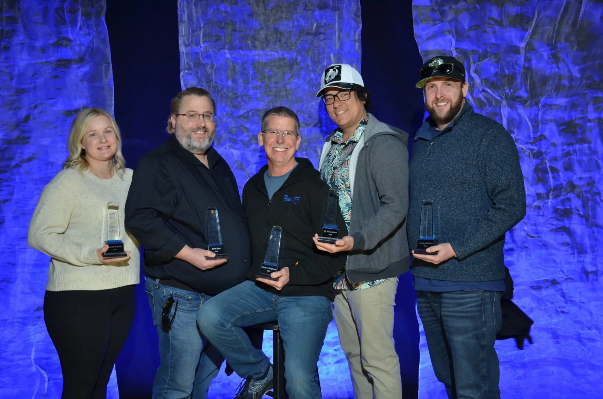 Group of five people posing with awards on stage at an event.