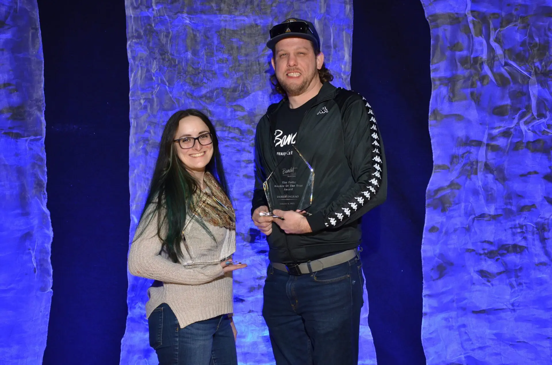 Two people posing together against a blue-lit stone wall, one holding an award.