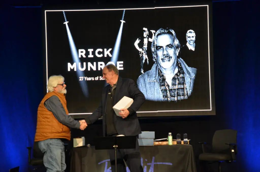 Two men shaking hands in front of a Rick Munroe tribute screen.