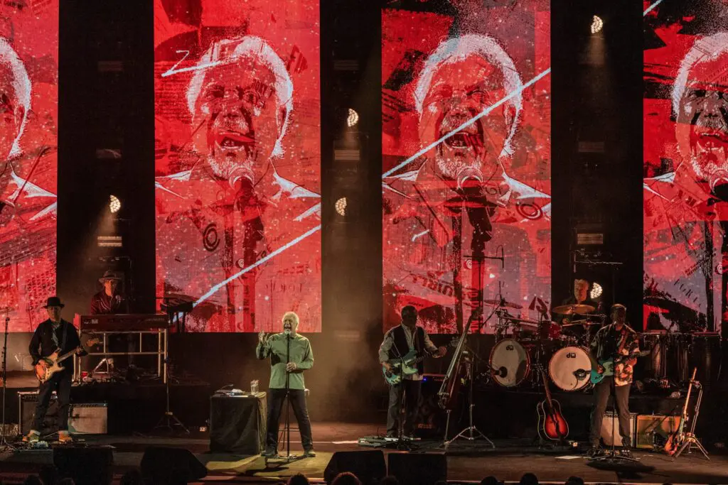 Band performs live on stage with large red visual screens behind.