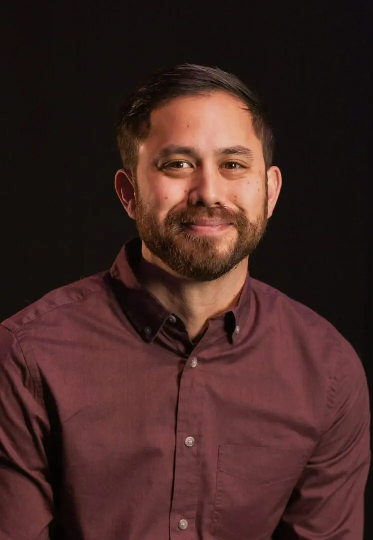 Portrait of a smiling man in a maroon shirt against a dark background.