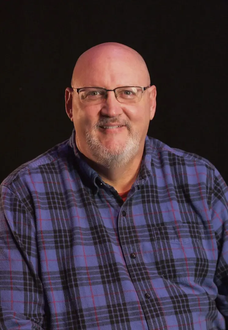 Smiling man with glasses and a plaid shirt against a dark background.