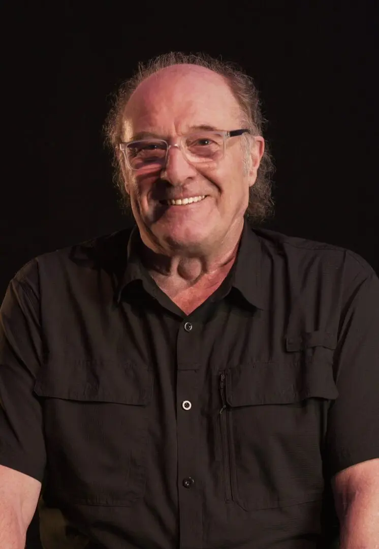 Smiling man with glasses wearing a black shirt against a dark background.