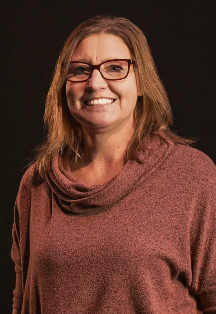 Smiling woman wearing glasses and a maroon sweater against a dark background.