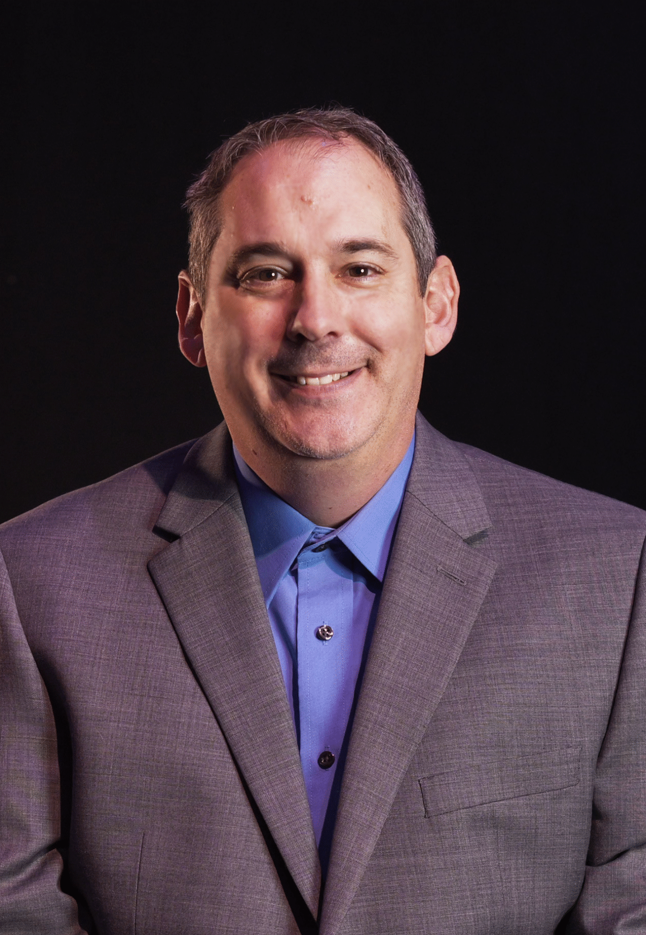 Smiling man in a grey suit and blue shirt against a black background.