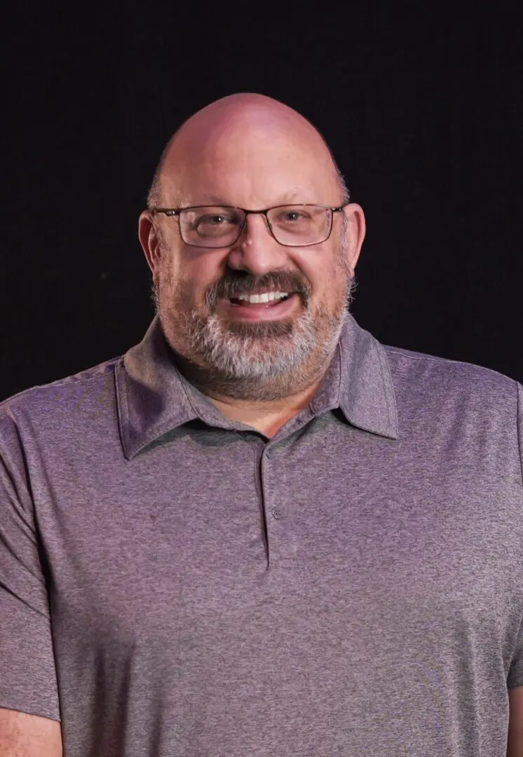 Smiling bald man with glasses and a gray polo shirt against a black background.