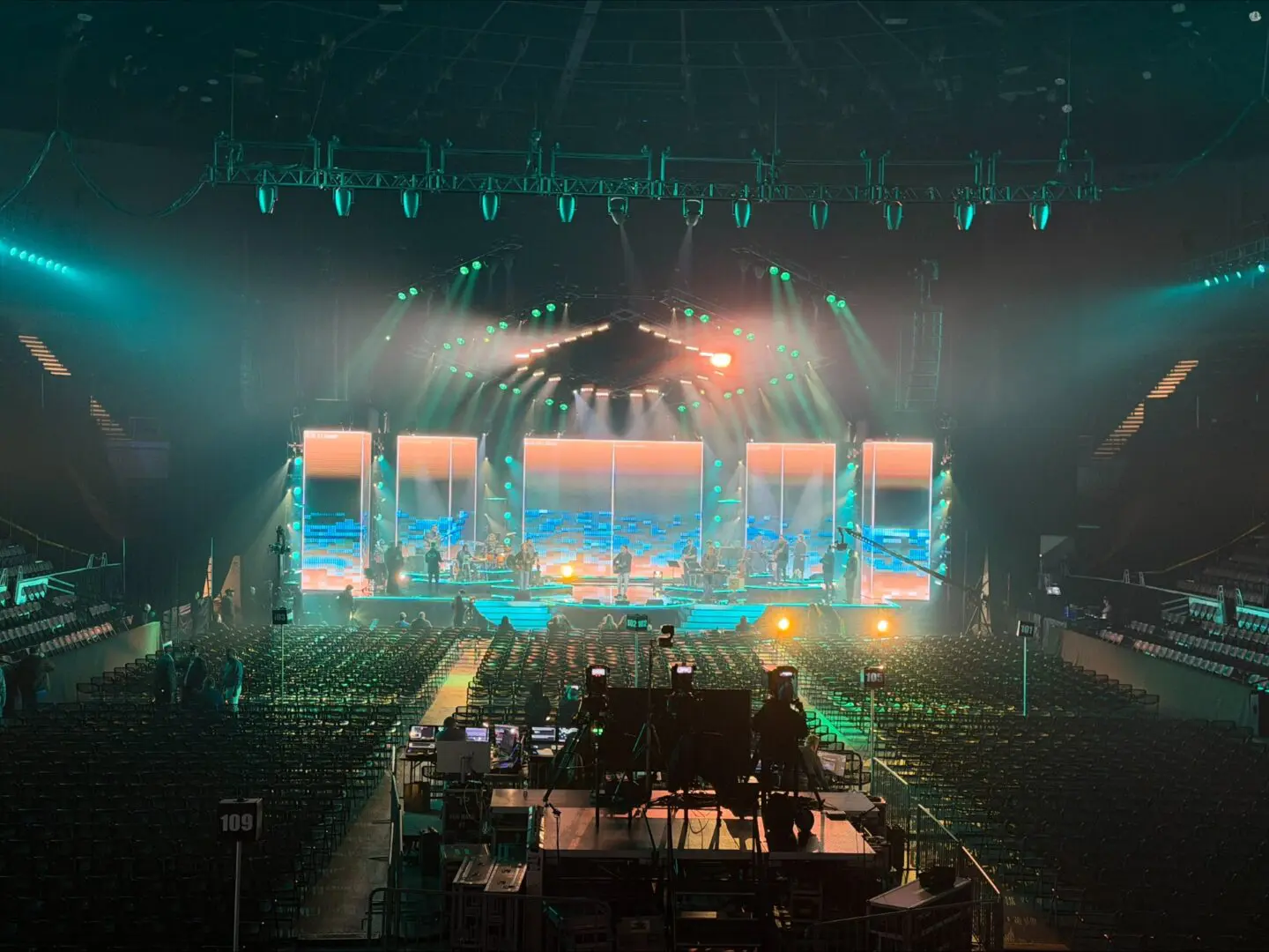 Concert stage illuminated with vibrant lights and large screens in a packed arena.