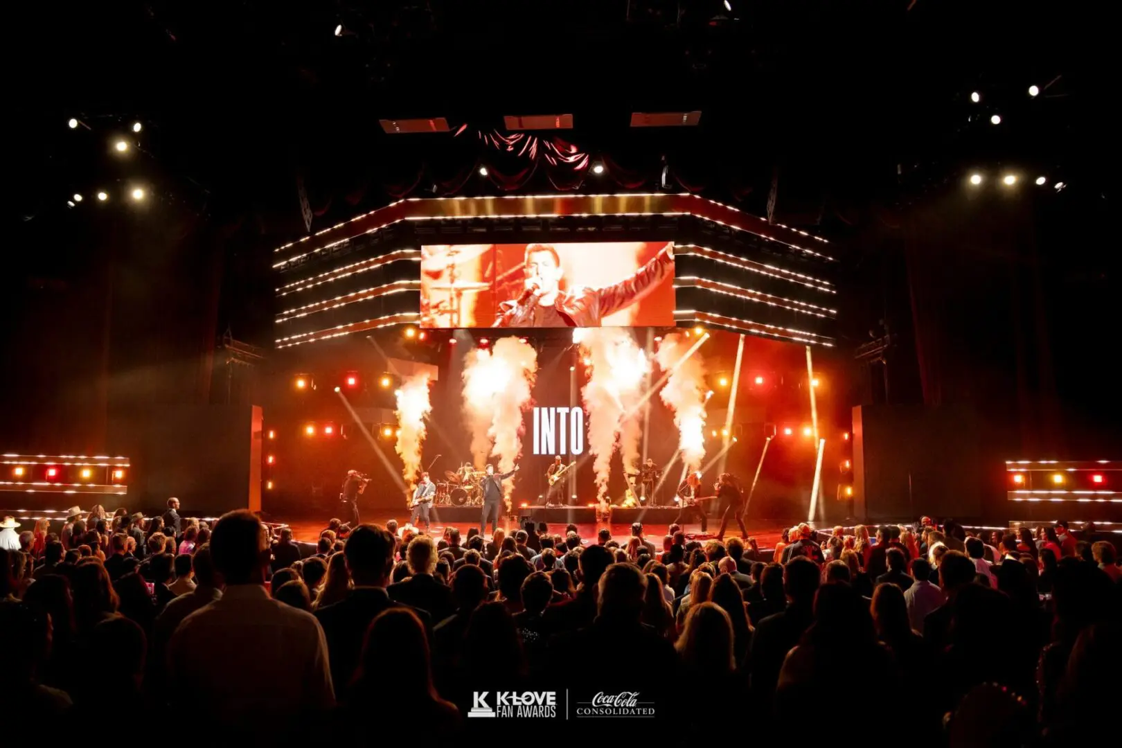 A nighttime concert with fiery stage effects and a large crowd.