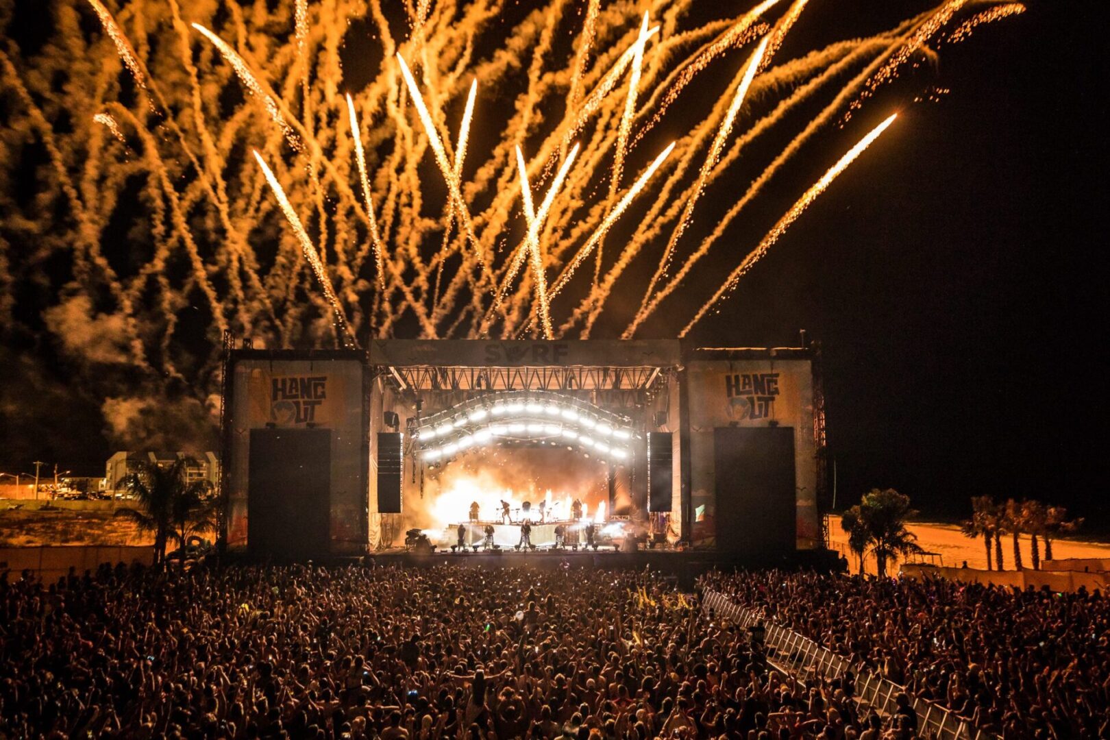 Fireworks light up the sky above a packed outdoor concert stage at night.