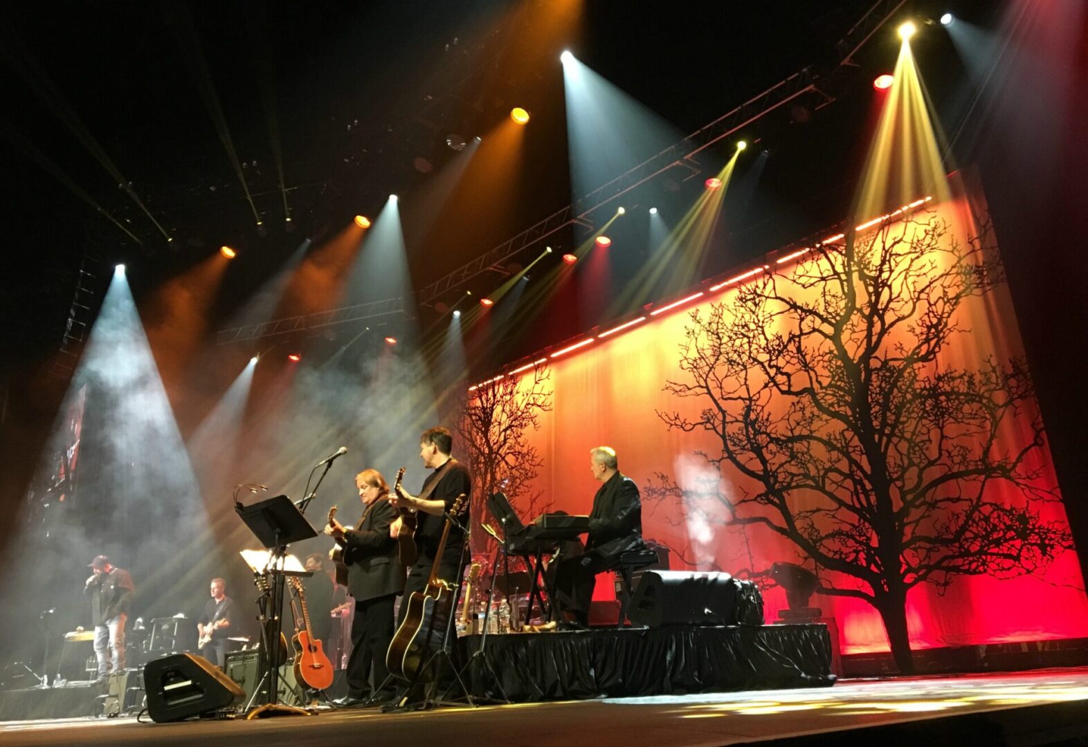 Musicians perform on stage with dramatic lighting and a tree backdrop.