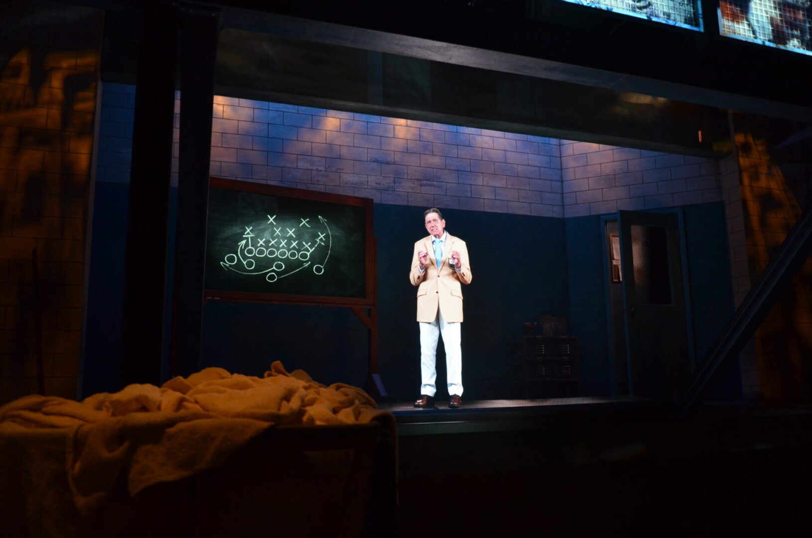 A man in white performs on a dimly lit stage with a chalkboard behind him.