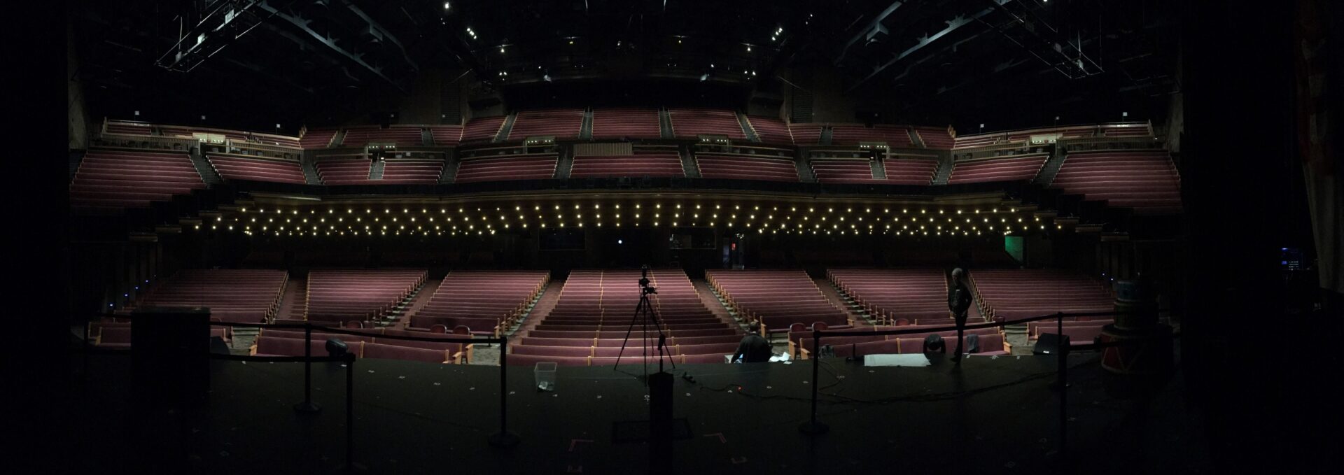 Empty theater stage with rows of red seats and dim lighting.