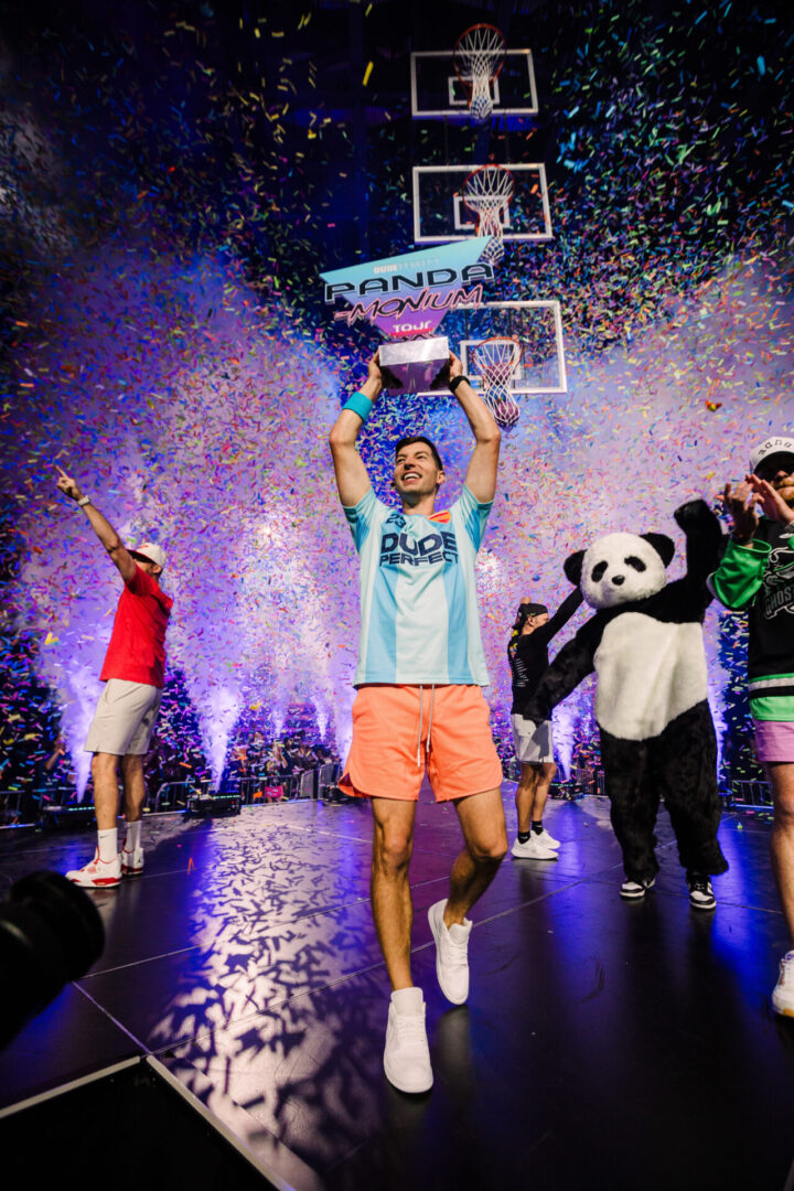 Excited man holding a trophy on a confetti-filled stage with people in costumes.