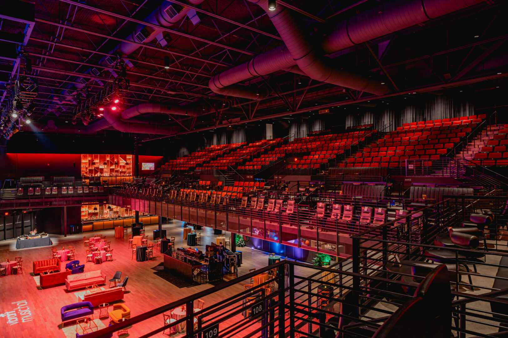 Empty arena with stage lighting and seating under purple and red lights.