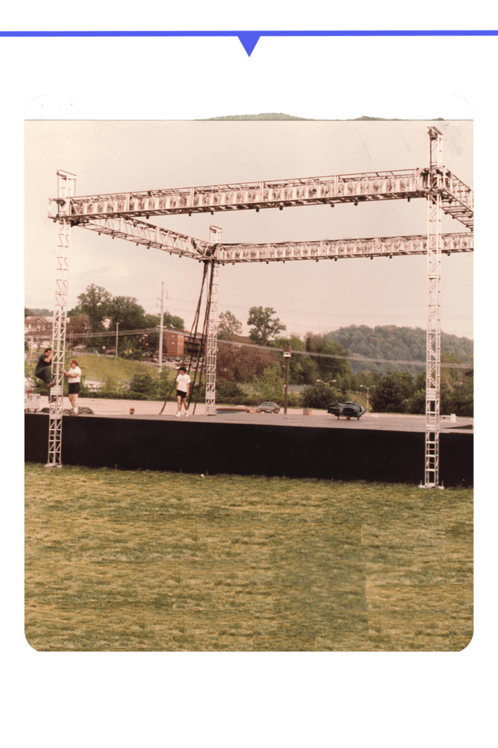 An outdoor stage setup with metal scaffolding and speakers on a grassy field.