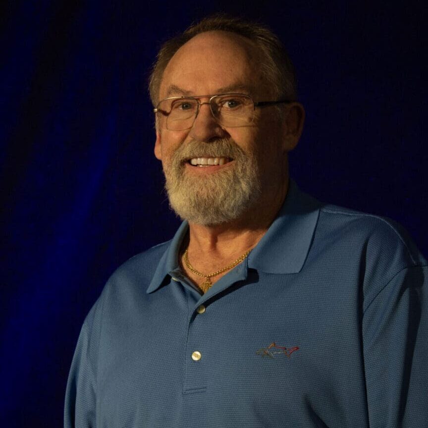 Smiling elderly man with glasses and beard in a blue polo shirt.