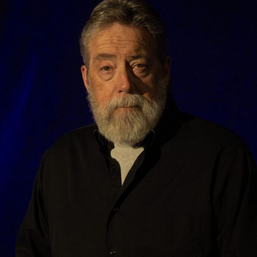 Serious elderly man with a beard against a dark blue background.