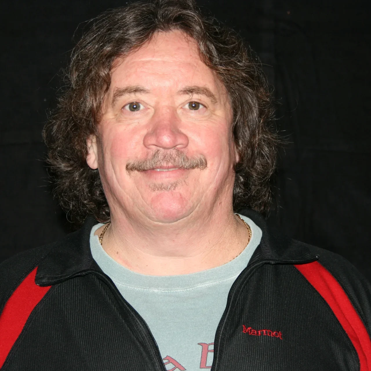 Smiling man with mustache and wavy hair in casual attire.