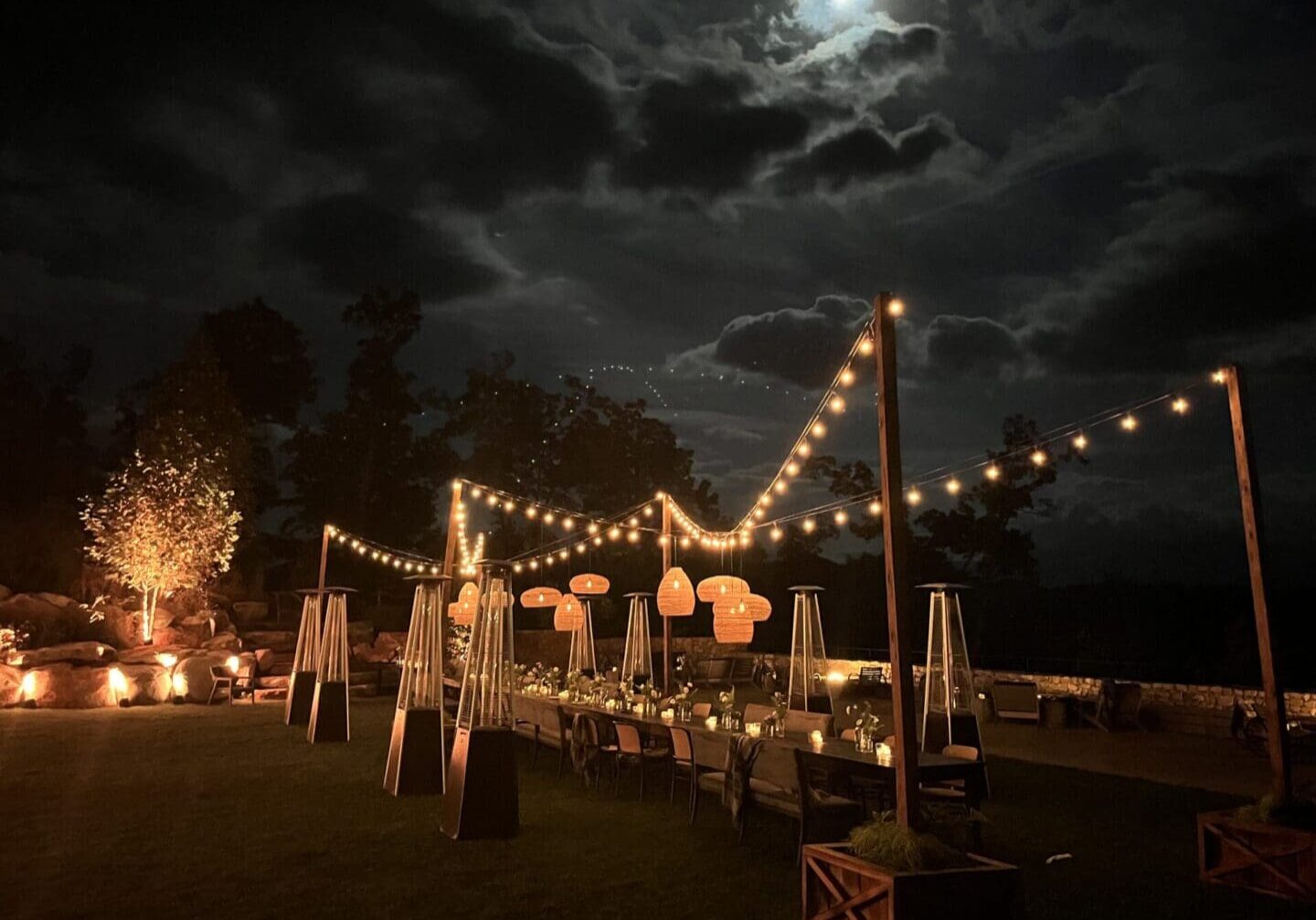 Outdoor dining setup under a moonlit sky with string lights.