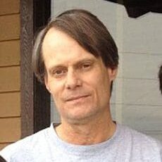 A middle-aged man with straight brown hair wearing a light gray shirt.