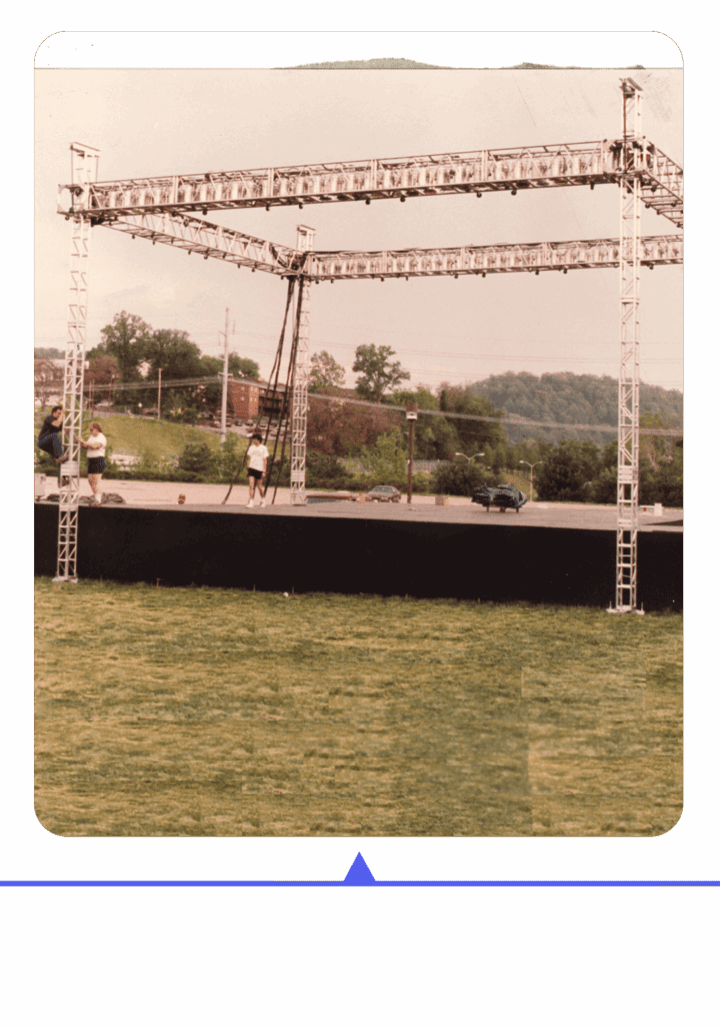 Outdoor stage setup with people preparing under a cloudy sky.
