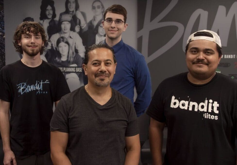 A group of five casually dressed men posing indoors in front of a photo wall.
