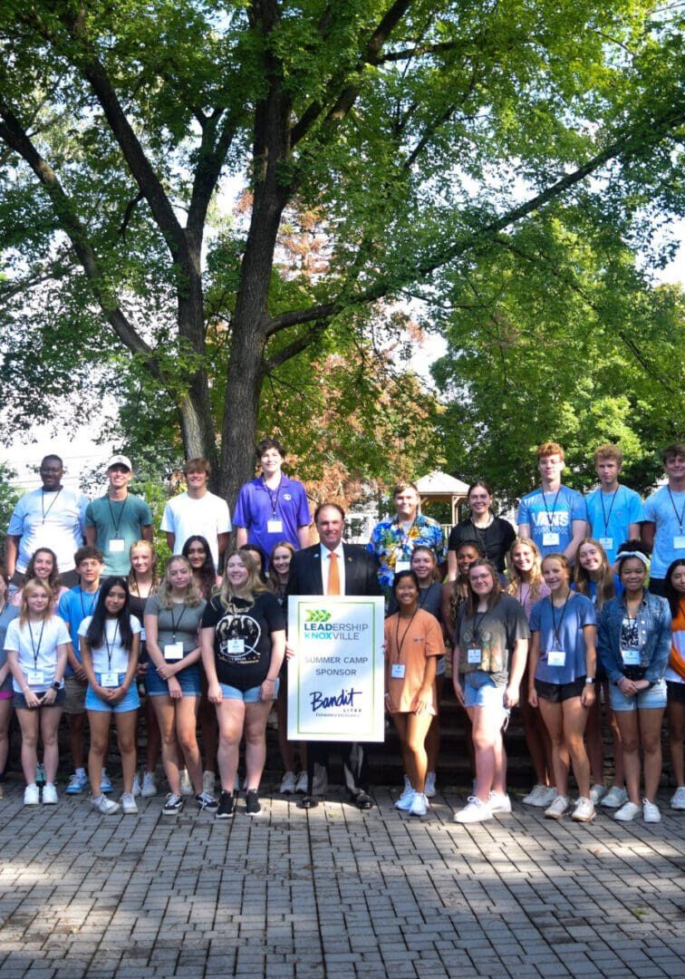 Large diverse group posing outdoors under trees with medals and a banner.